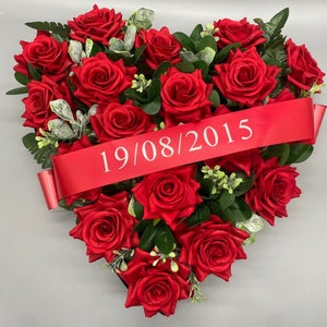 May include: A heart-shaped floral arrangement composed of vibrant red roses, green leaves, and a red ribbon. The ribbon displays the date "19/08/2015" in white text. The arrangement is a memorial or decorative piece.