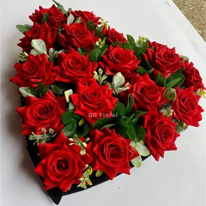 May include: A heart-shaped floral arrangement filled with vibrant red roses and green foliage. The roses are densely packed, creating a full and lush appearance. The arrangement is set in a black container, with the text "GG Floral" visible.