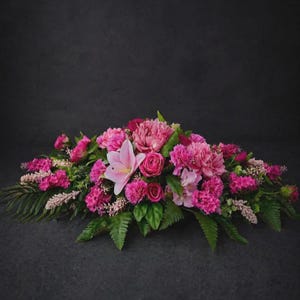 May include: A horizontal floral arrangement featuring a mix of vibrant pink carnations, roses, and peonies, accented by a single pale pink lily. The bouquet includes various green fern fronds and small, light-coloured filler flowers throughout.
