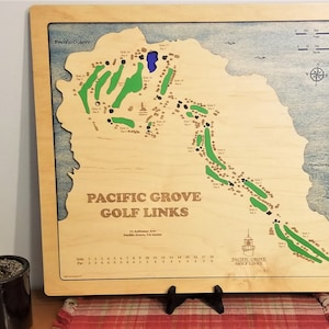 May include: A wooden map of the Pacific Grove Golf Links, showing the layout of the course with green and brown details. The map is framed and includes a scorecard with the par for each hole. The text "PACIFIC GROVE GOLF LINKS" is printed on the map.