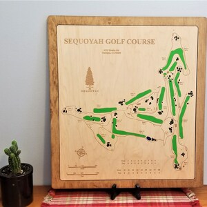 May include: A wooden plaque with a map of the Sequoyah Golf Course in Oakland, California. The map shows the layout of the 18 holes, with green fairways and black sand traps. The plaque also includes the address of the golf course: 4550 Healy Rd, Oakland, CA 94605.