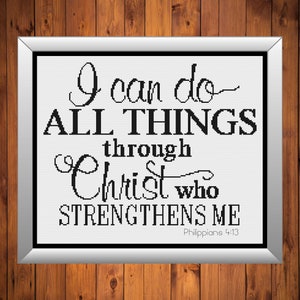 May include: A framed cross-stitch artwork with the text "I can do ALL THINGS through Christ who STRENGTHENS ME" in black on a white background. The frame is silver, and the background is a wooden surface.