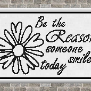 May include: A framed cross-stitch artwork with a daisy and the text "Be the Reason someone smiles today" in black on a white background. The frame is silver, and the background is a brick wall.