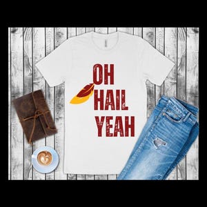 May include: White t-shirt with the text "OH HAIL YEAH" in burgundy, featuring a feather graphic. The shirt is paired with blue jeans, a leather-bound journal, and a cup of coffee with latte art.