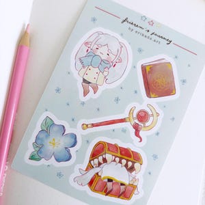 Op de afbeelding: Een stickervel met illustraties van "Frieren's Journey" door erikads.art. Het vel bevat een personage, een boek, een staf, een schatkist en een blauwe bloem. De achtergrond is lichtblauw met kleine bloem- en sterontwerpen.