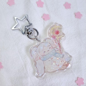 May include: A clear acrylic keychain with a cartoon character with long white hair, a blue scarf, and a staff. The keychain has a silver star-shaped clasp. Pink flower-shaped confetti is scattered around the keychain.