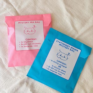 Op de afbeelding: Twee kleurrijke mystery bags. Eén is roze en de andere is blauw. Op de roze tas staat "Mystery Mix Bag" en bevat een sleutelhanger, gestanste stickers en een verrassingsartikel. De blauwe tas zegt "Stationery Mania Mystery Bag" en bevat een plaknotitieblok of stickerblad, gestanste stickers en een verrassingsartikel.