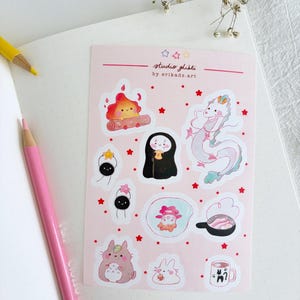 Op de afbeelding: Een vel roze stickers met Studio Ghibli-personages, waaronder een kampvuur, een draak en een roetgeest. De stickers hebben een witte rand en zijn omgeven door rode sterren. De tekst "studio ghibli by erikads.art" staat bovenaan.