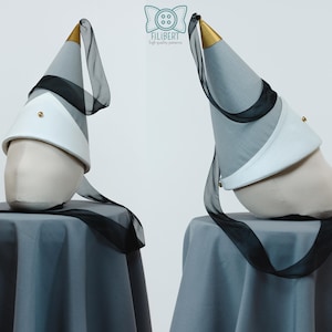 May include: A conical hat in grey fabric with a white brim and a gold-colored tip. The hat features a long, sheer black ribbon attached to the top and a small gold stud on the white brim. The logo Filibert high quality patterns is visible.