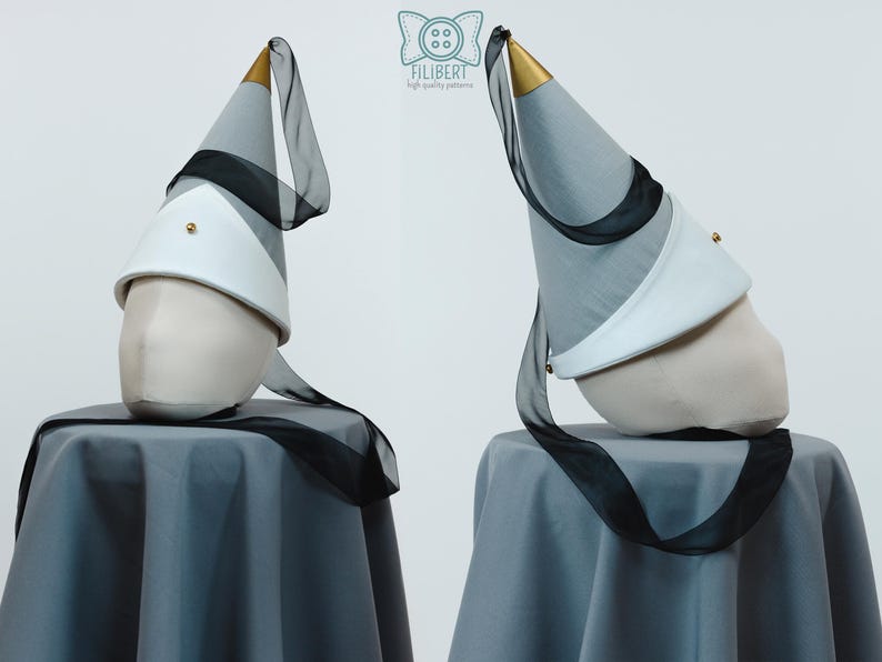 May include: A conical hat in grey fabric with a white brim and a gold-colored tip. The hat features a long, sheer black ribbon attached to the top and a small gold stud on the white brim. The logo Filibert high quality patterns is visible.