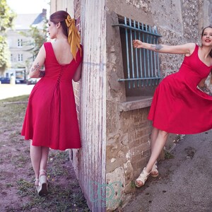 May include: Two women wearing red dresses pose outdoors. The dresses have a scoop neckline and a flared skirt. One dress has a lace-up back. Both women wear sandals and have their hair up with a yellow bow.