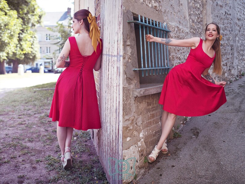 May include: Two women wearing red dresses pose outdoors. The dresses have a scoop neckline and a flared skirt. One dress has a lace-up back. Both women wear sandals and have their hair up with a yellow bow.