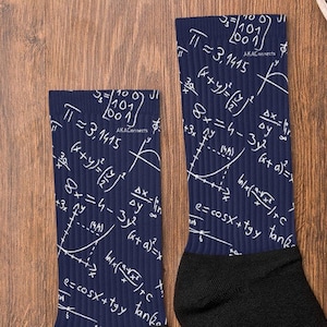 May include: A pair of black and navy blue socks with a mathematical formula pattern. The socks feature white equations and symbols, including "π = 3.1415". The socks are displayed on a wooden surface with a pair of gray sneakers.
