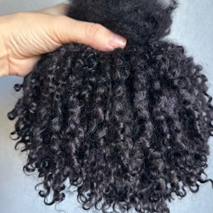 May include: A hair extension piece featuring tightly coiled, dark black curls. The texture is dense and voluminous, with individual ringlets cascading downward from a gathered top section, creating a rounded, full shape with a natural appearance.