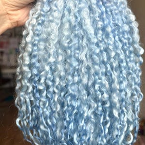 May include: A bundle of light blue curly hair extensions, held together with a red ribbon. The hair has a soft, fluffy texture and a mix of light and medium blue tones, creating a natural look.