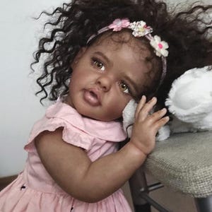 Peut inclure: Une poup&eacute;e r&eacute;aliste &agrave; la peau fonc&eacute;e, aux yeux marron et aux longs cheveux noirs boucl&eacute;s. La poup&eacute;e porte une robe rose clair avec un petit col et un motif de minuscules papillons sombres, agr&eacute;ment&eacute;e d'un bandeau floral &agrave; fleurs blanches et roses.