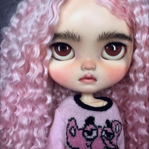 Peut inclure: Une poup&eacute;e aux longs cheveux boucl&eacute;s rose clair, aux yeux marron fonc&eacute; et aux sourcils &eacute;pais. La poup&eacute;e porte un pull &agrave; manches longues rose clair avec une impression graphique d'un personnage de dessin anim&eacute; rose aux contours noirs.