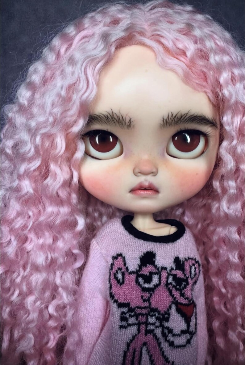 Peut inclure: Une poup&eacute;e aux longs cheveux boucl&eacute;s rose clair, aux yeux marron fonc&eacute; et aux sourcils &eacute;pais. La poup&eacute;e porte un pull &agrave; manches longues rose clair avec une impression graphique d'un personnage de dessin anim&eacute; rose aux contours noirs.