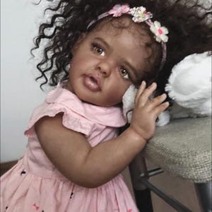 May include: A realistic baby doll with dark curly hair, brown eyes, and a floral headband. The doll wears a light pink dress with a small butterfly print and a collar. It has a soft, lifelike complexion and is positioned with one arm resting.