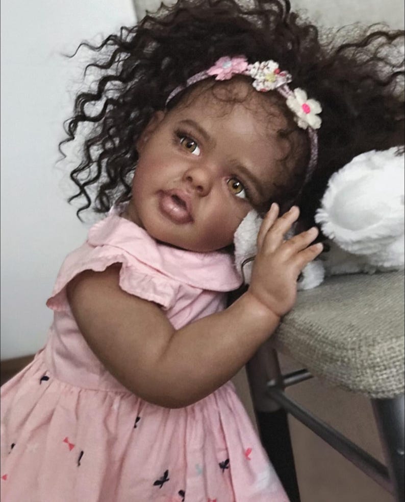 May include: A realistic baby doll with dark curly hair, brown eyes, and a floral headband. The doll wears a light pink dress with a small butterfly print and a collar. It has a soft, lifelike complexion and is positioned with one arm resting.
