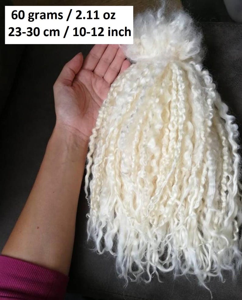 Peut inclure: Un lot de m&egrave;ches de laine blanc cass&eacute;, longues et boucl&eacute;es. Le texte sur l'image indique un poids de 60 grammes et une longueur de 23 &agrave; 30 cm. Les fibres pr&eacute;sentent une texture ondul&eacute;e et sont dispos&eacute;es en un faisceau vertical dense.
