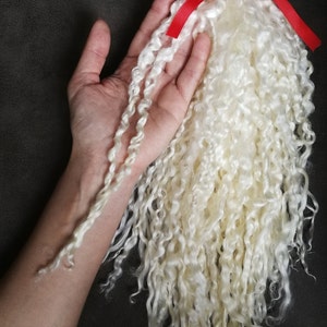 May include: A bundle of off-white, curly, long-staple fiber weighing 60 grams or 2.11 ounces, secured at the top with a bright red ribbon. The texture is fine and ringlet-like, showcasing a natural, soft appearance for crafting and textile projects.