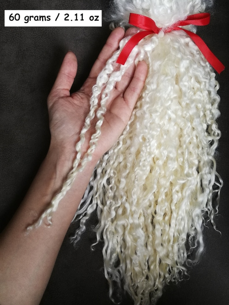 May include: A bundle of off-white, curly, long-staple fiber weighing 60 grams or 2.11 ounces, secured at the top with a bright red ribbon. The texture is fine and ringlet-like, showcasing a natural, soft appearance for crafting and textile projects.