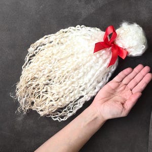 May include: A bundle of off-white, curly hair tied with a bright red ribbon. The hair is arranged on a dark grey surface, with a hand visible in the frame. The hair has a natural, textured appearance.
