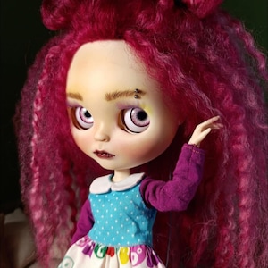 May include: A doll with vibrant magenta curly hair styled in two top knots. The doll has pale skin, light purple eyes, and a small silver eyebrow piercing. It wears a blue polka-dot top with long purple sleeves and a white skirt featuring colorful bear prints.