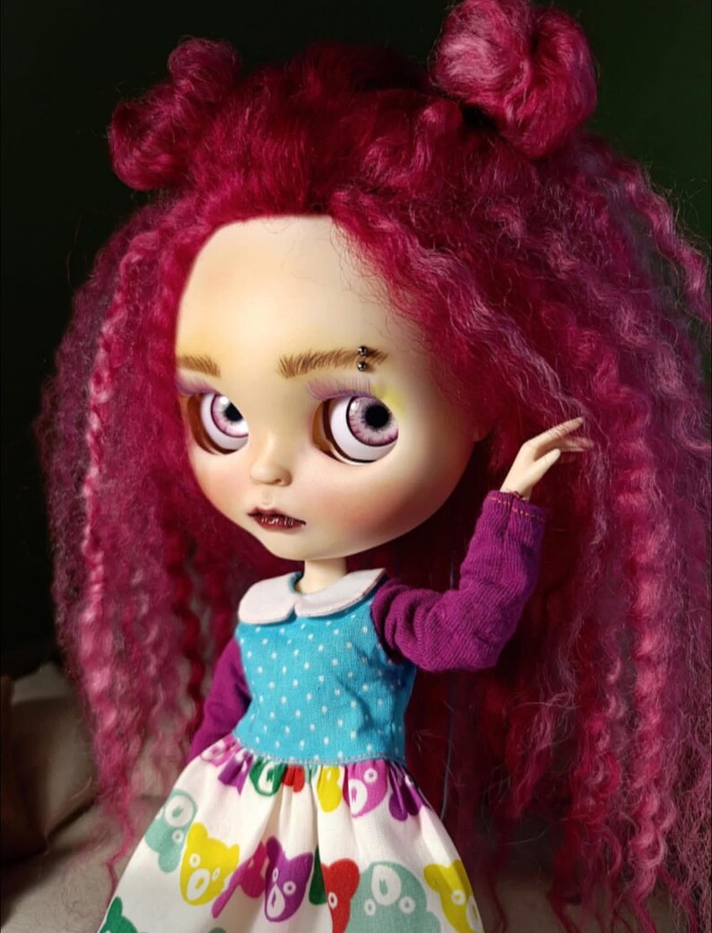 May include: A doll with vibrant magenta curly hair styled in two top knots. The doll has pale skin, light purple eyes, and a small silver eyebrow piercing. It wears a blue polka-dot top with long purple sleeves and a white skirt featuring colorful bear prints.