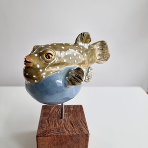 Może przedstawiać: Ceramiczna rzeźba ryby rozdymki z brązowym grzbietem w białe kropki i gładkim niebieskim brzuchem. Ryba jest osadzona na cienkim metalowym pręcie przymocowanym do kwadratowej, ciemnobrązowej drewnianej podstawy, tworząc dekoracyjny element wystroju.