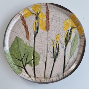 May include: A round ceramic plate with a cream-coloured crackle glaze finish. The surface features pressed botanical elements including yellow flowers, green leaves, and brown stalks arranged in a vertical pattern across the centre of the dish.
