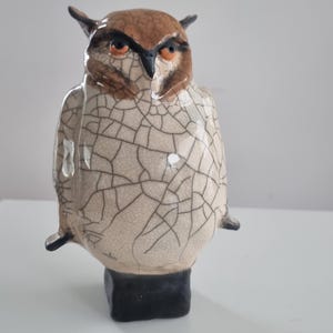 Może przedstawiać: Ceramiczna rzeźba sowy z pękniętym szkliwem. Sowa jest brązowa i biała z czarnymi oczami i czarnym dziobem. Sowa stoi na czarnej podstawie.