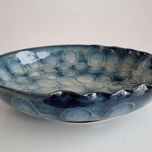 May include: A handmade ceramic bowl with a unique, textured surface. The bowl features a deep blue glaze with lighter blue and beige accents, and a wavy, scalloped rim. This decorative bowl is perfect for serving or display.