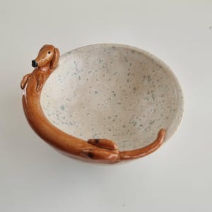 Op de afbeelding: Een keramische kom met een teckelhond ontwerp. De kom is gebroken wit met blauwe spikkels en de hond is bruin met zwarte accenten. De hond wikkelt zich om de rand van de kom en creëert een uniek decoratief stuk.