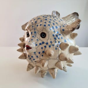 Peut inclure: Une figurine de poisson-globe en céramique avec un corps couleur crème doté d'une finition émaillée craquelée, orné de petits pois bleus. La sculpture présente des épines coniques saillantes, des yeux sombres et une petite bouche rouge.