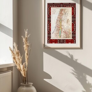 May include: A framed vintage map of Palestine with a red and white patterned border. The map is in a light wood frame and is hanging on a white wall.