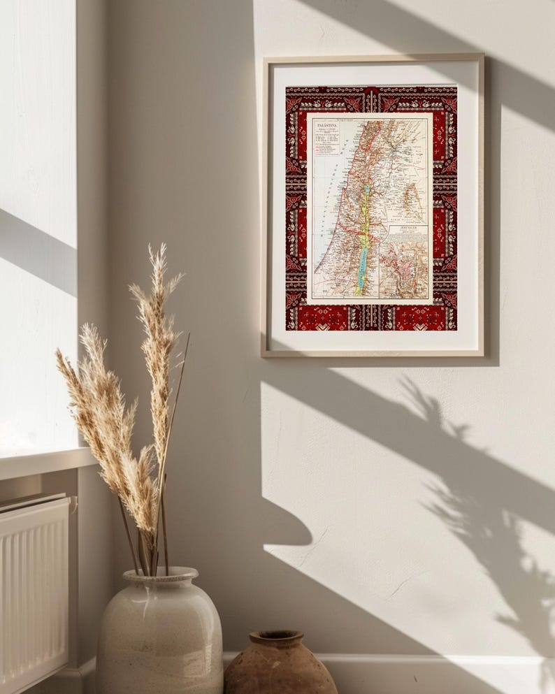 May include: A framed vintage map of Palestine with a red and white patterned border. The map is in a light wood frame and is hanging on a white wall.