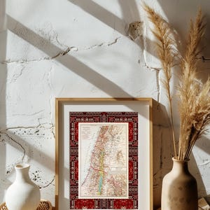 May include: A framed vintage map of Palestine with a red and white patterned border. The map is displayed on a white wall with a vase of dried pampas grass on the right side of the frame.