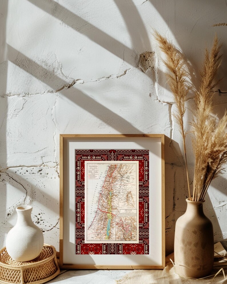 May include: A framed vintage map of Palestine with a red and white patterned border. The map is displayed on a white wall with a vase of dried pampas grass on the right side of the frame.