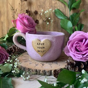 May include: Purple ceramic mug with a heart-shaped design that says "Love Your Self". The mug is sitting on a wooden slice with flowers and greenery around it.