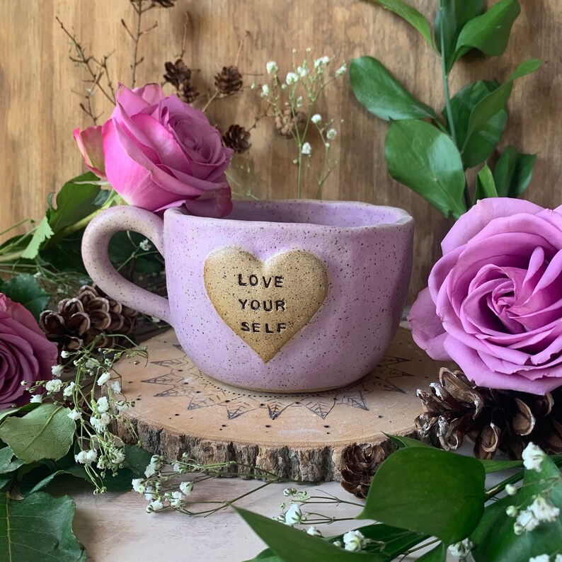 May include: Purple ceramic mug with a heart-shaped design that says "Love Your Self". The mug is sitting on a wooden slice with flowers and greenery around it.