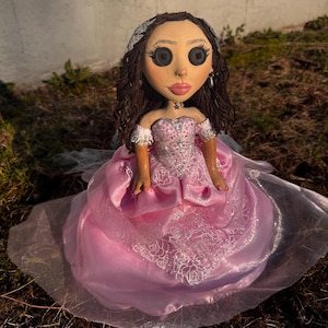 Peut inclure: Une poup&eacute;e aux cheveux bruns boucl&eacute;s, yeux en boutons noirs et l&egrave;vres roses, v&ecirc;tue d'une robe de bal rose &agrave; &eacute;paules d&eacute;nud&eacute;es avec des superpositions de dentelle blanche, des ornements argent&eacute;s et un diad&egrave;me argent&eacute;, avec un collier &eacute;toile.
