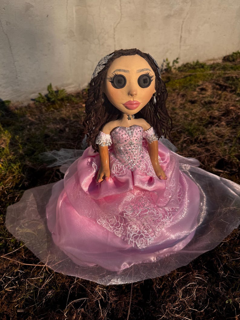 Peut inclure: Une poup&eacute;e aux cheveux bruns boucl&eacute;s, yeux en boutons noirs et l&egrave;vres roses, v&ecirc;tue d'une robe de bal rose &agrave; &eacute;paules d&eacute;nud&eacute;es avec des superpositions de dentelle blanche, des ornements argent&eacute;s et un diad&egrave;me argent&eacute;, avec un collier &eacute;toile.