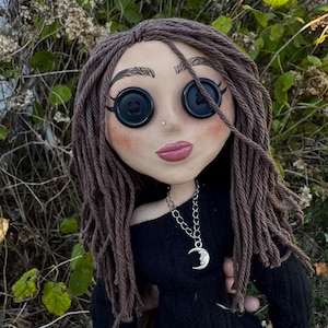 Peut inclure: Une poup&eacute;e avec des cheveux en laine brune, des yeux en boutons noirs et des l&egrave;vres roses. La poup&eacute;e porte un haut noir &agrave; manches longues et un collier &agrave; cha&icirc;ne argent&eacute;e avec un pendentif en forme de croissant de lune avec un visage.