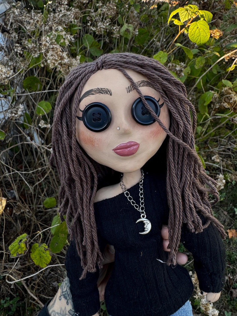 Peut inclure: Une poup&eacute;e avec des cheveux en laine brune, des yeux en boutons noirs et des l&egrave;vres roses. La poup&eacute;e porte un haut noir &agrave; manches longues et un collier &agrave; cha&icirc;ne argent&eacute;e avec un pendentif en forme de croissant de lune avec un visage.