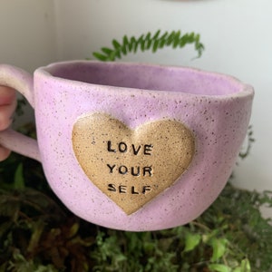 May include: A light purple ceramic mug with a speckled finish. The mug has a heart-shaped handle and a gold heart on the front that says "LOVE YOUR SELF".