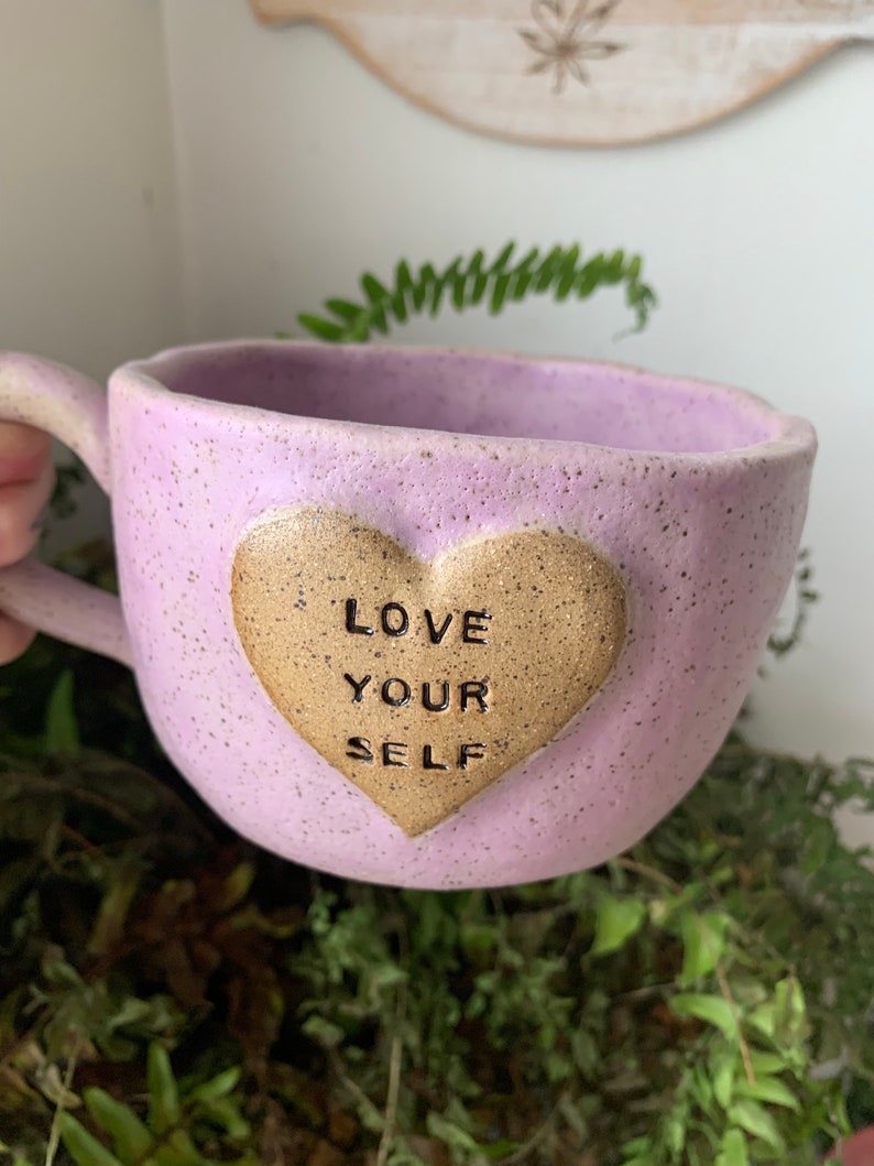 May include: A light purple ceramic mug with a speckled finish. The mug has a heart-shaped handle and a gold heart on the front that says "LOVE YOUR SELF".