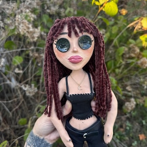 Peut inclure: Une poup&eacute;e avec de longs cheveux en laine brun fonc&eacute;, de grands yeux en boutons noirs et des traits du visage peints. La poup&eacute;e porte un haut court noir avec des coutures blanches, un pantalon noir avec une ceinture et un collier dor&eacute; avec un croissant de lune.