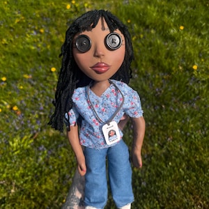 Peut inclure: Une poup&eacute;e &agrave; la peau fonc&eacute;e, aux cheveux en laine noire et aux yeux en boutons noirs. Elle porte un chemisier bleu clair &agrave; manches courtes avec un motif floral rose et blanc, un pantalon en jean bleu et des chaussures blanches. Un badge Brandi.
